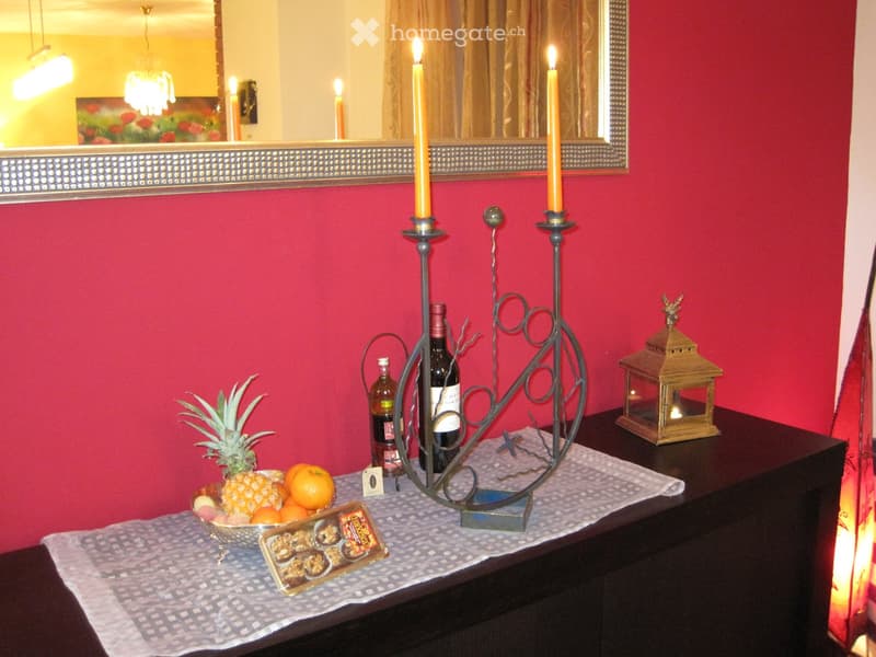 A small table with a decorative arrangement including a bowl of fruit, wine bottles, candles, a lantern, and a cake, set against a red wall with a mirror.
