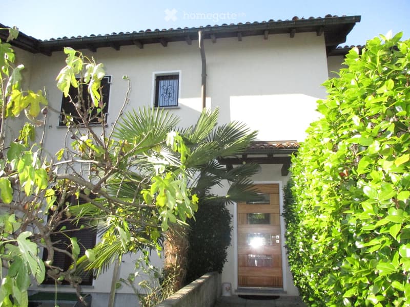 Two-story house with beige stucco walls, wooden front door, and decorative window grilles, surrounded by lush greenery and palm trees.