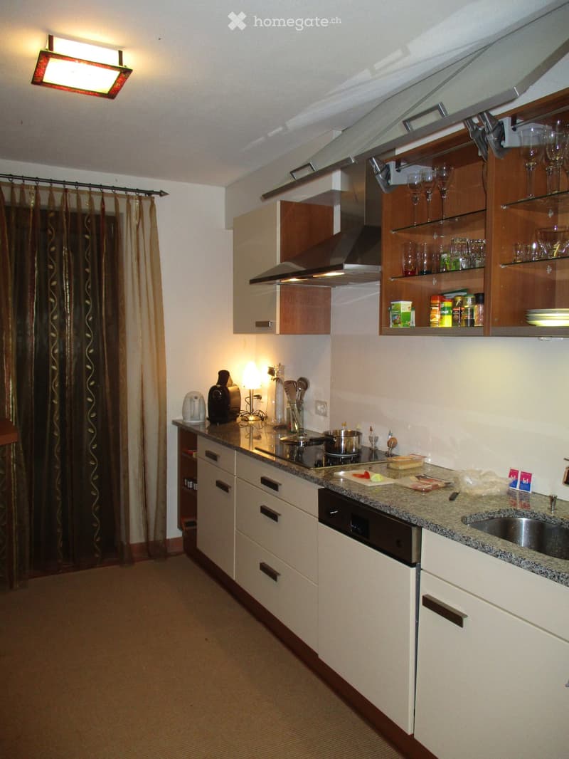 Modern kitchen with white cabinets, granite countertop, built-in oven, and various appliances. Glassware and dishes are displayed on open shelves above the counter.