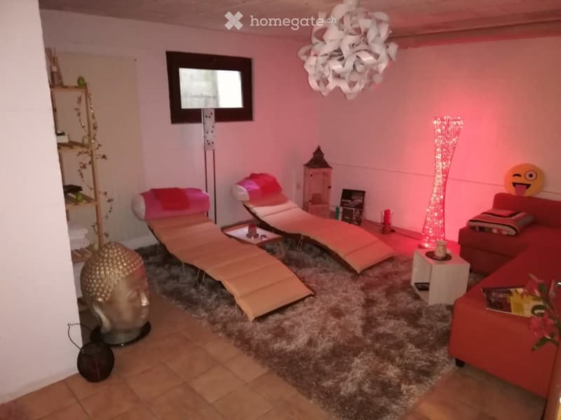 A modern, well-lit room with a red sofa, two sun loungers, a decorative floor lamp, a large mirror, a sculpture, and a small table with books and flowers.