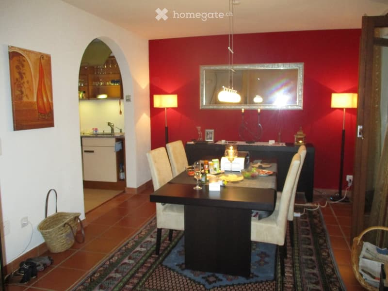 Dining room with a dark wooden dining table surrounded by white chairs, a red accent wall, a large mirror, floor lamps, and an open kitchen area in the background.