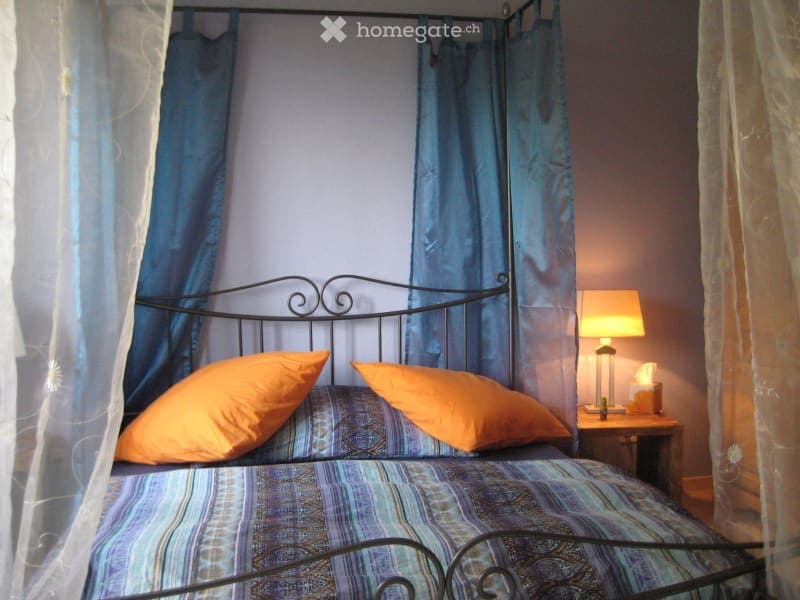 Cozy bedroom with a metal-framed bed, blue and orange bedding, a nightstand with a lamp, and blue curtains.