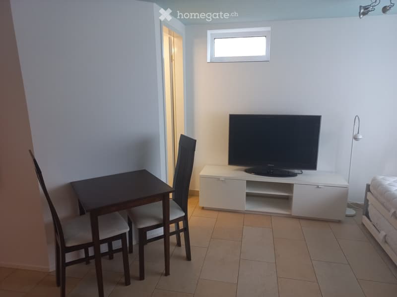 A modern, minimalist living space with a TV on a white stand, a small dining table with two chairs, and a floor lamp near the TV.