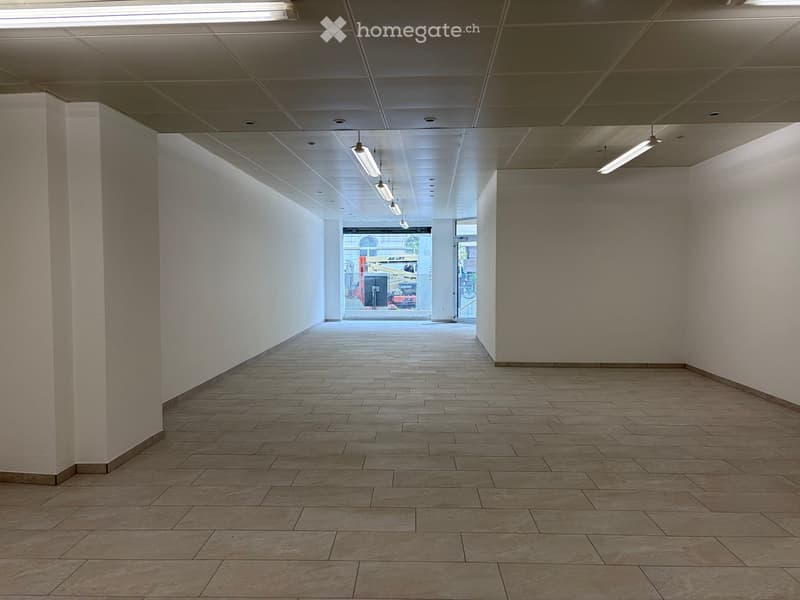 Empty hallway with tiled floor, white walls, and fluorescent lighting. Features a large window at the end and a pillar on the left side.