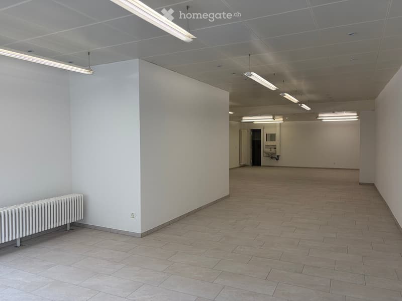 Empty room with white walls, tiled floor, and a radiator on one wall. The space is well-lit with ceiling lights and has a doorway leading to another area.
