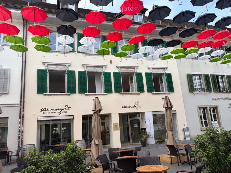 Facade of a two-story building with green shutters, multiple windows, and colorful umbrellas hanging above. The ground floor features a café with outdoor seating, tables, and chairs.