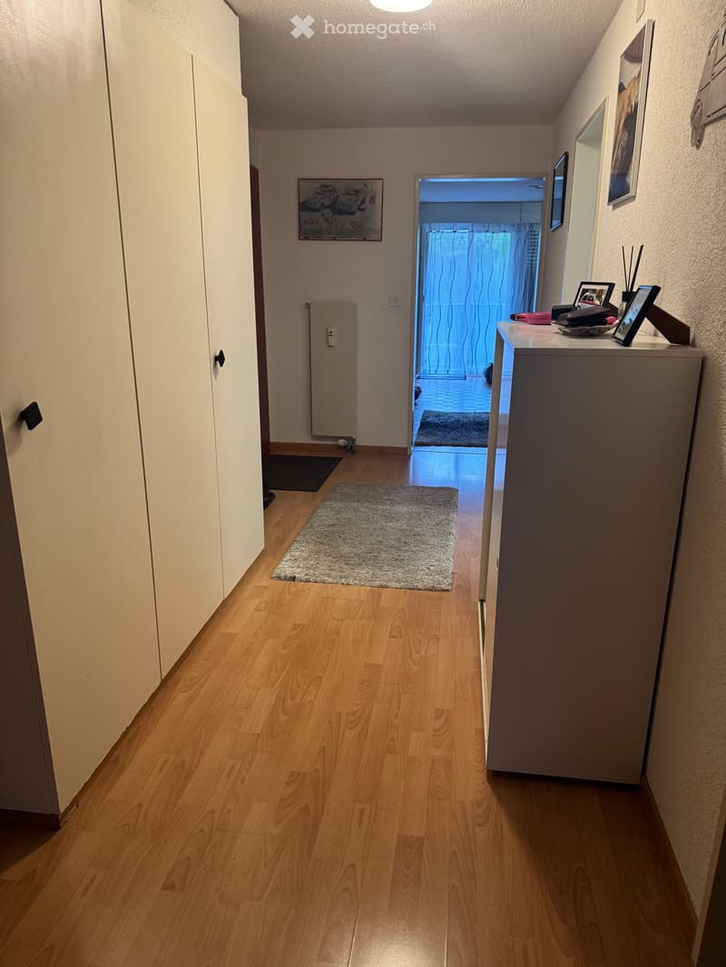 A narrow hallway with wooden flooring, white walls, and a small rug in the center. A small table with decorative items is visible on the right side, and a doorway leads to another room with blue lighting.