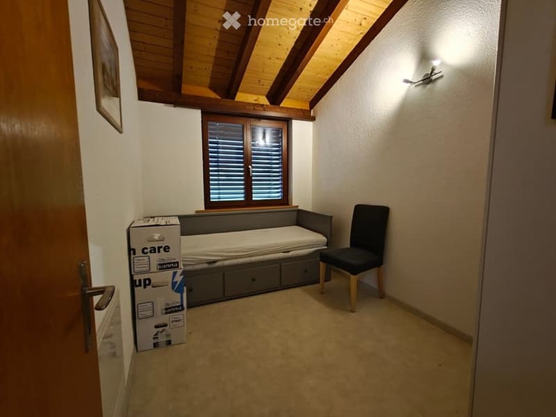 Cozy attic bedroom with wooden slanted ceilings, a window with shutters, a sofa bed, a chair, and a storage unit. A cardboard box is placed near the door.