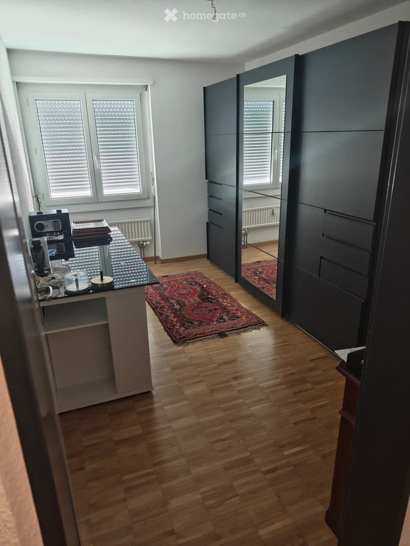 A furnished room with wooden flooring, a large mirror cabinet, a small table with various items, and a patterned rug.
