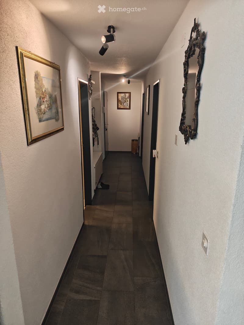 A narrow hallway with tiled flooring, white walls, and several doors leading to different rooms. There are framed pictures on the walls and a small bench with a shoe placed on it.