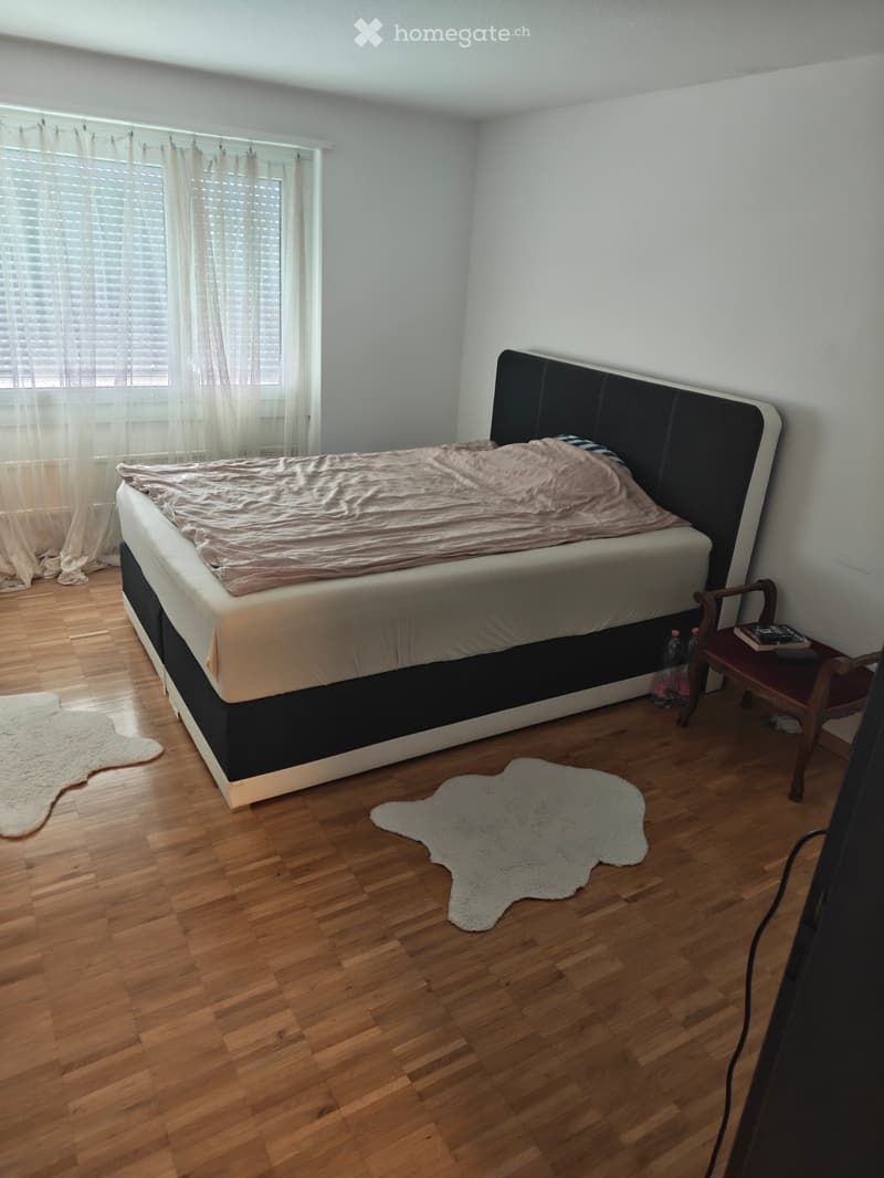 A well-lit bedroom with a large bed, a wooden chair, a small table, and two white rugs on the floor.