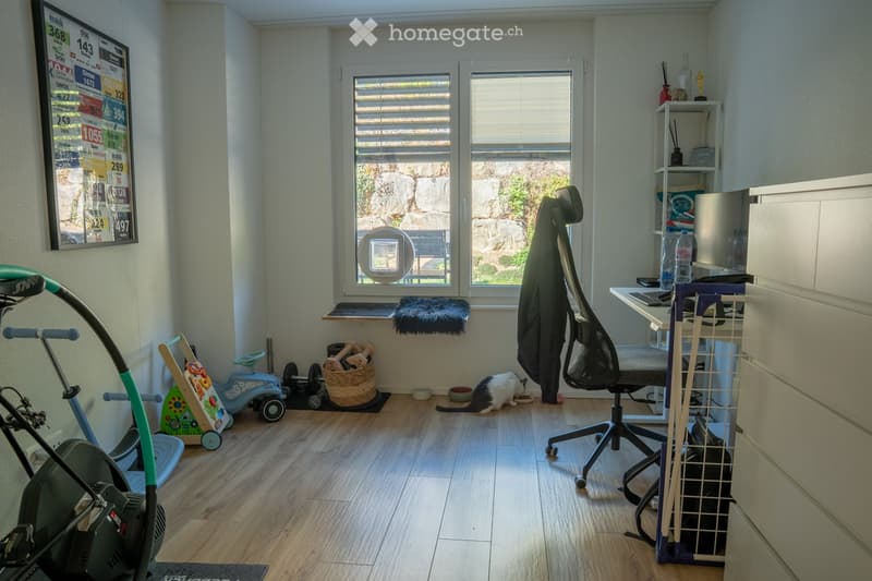 A multi-purpose room with a desk, exercise bike, cat area, and children's toys. Features a large window with blinds, a framed poster on the wall, and a small shelf with various items.