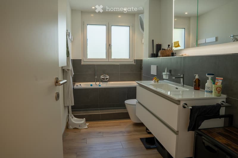 Modern bathroom with a bathtub, sink, toilet, and large mirror. Features dark tiled walls, wooden flooring, and various toiletries on the sink counter.