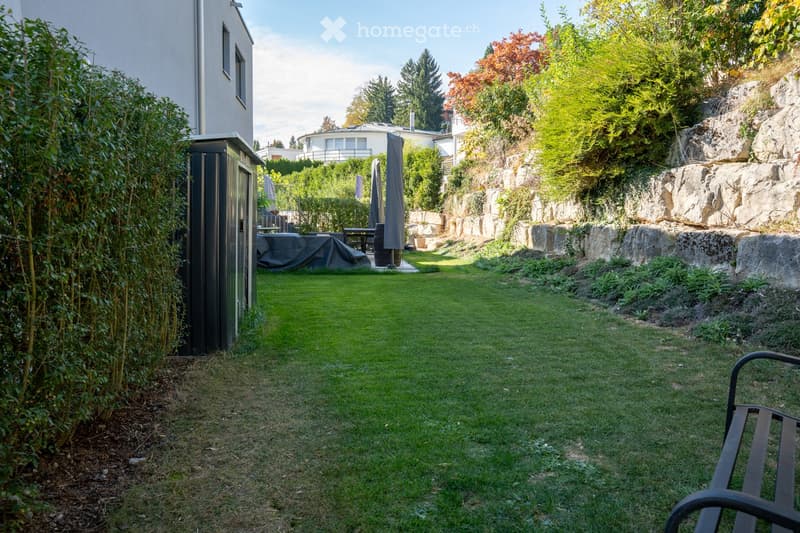 Well-maintained garden with a lawn, stone wall, covered patio area, and surrounding greenery.