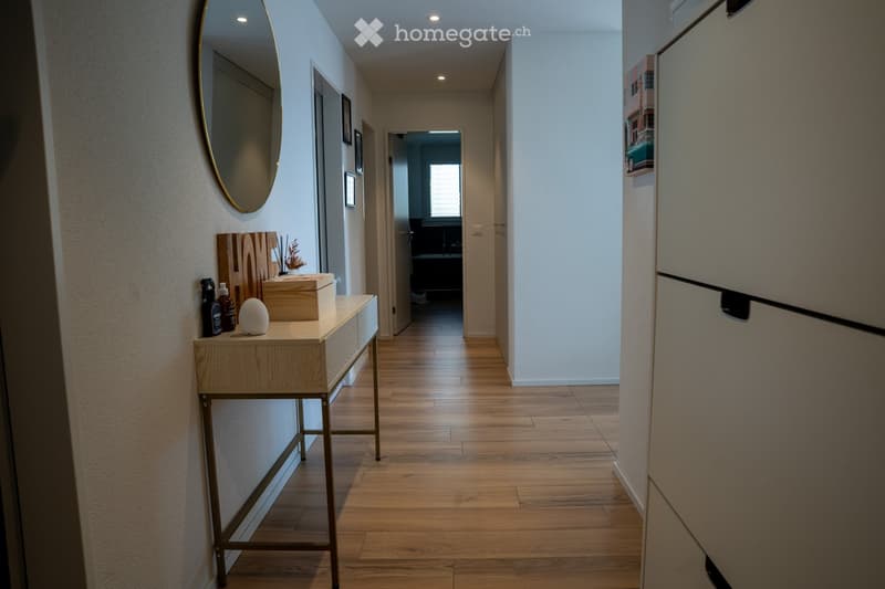 Modern hallway with wooden flooring, a small console table with decorative items, a round mirror on the wall, and a view into an adjacent room with a window.
