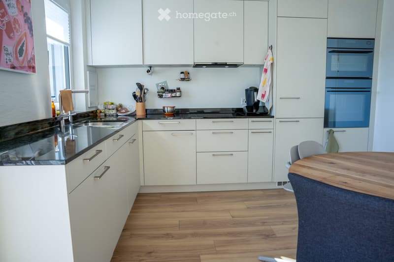 Modern kitchen with white cabinets, wooden flooring, and a central island. Features a sink, oven, and a small dining area with a table and chairs.