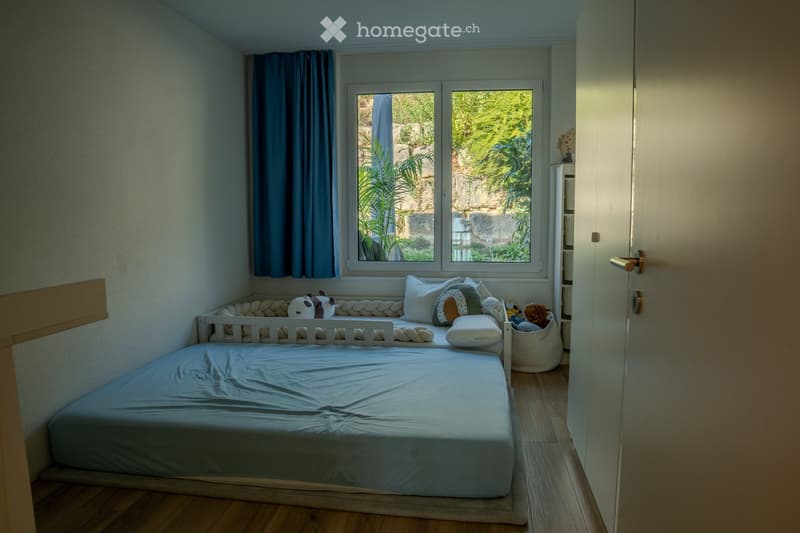 Cozy bedroom with a single bed, pillows, a curtain, a toy basket, and a wardrobe. The room has wooden flooring and a window with a view of greenery.