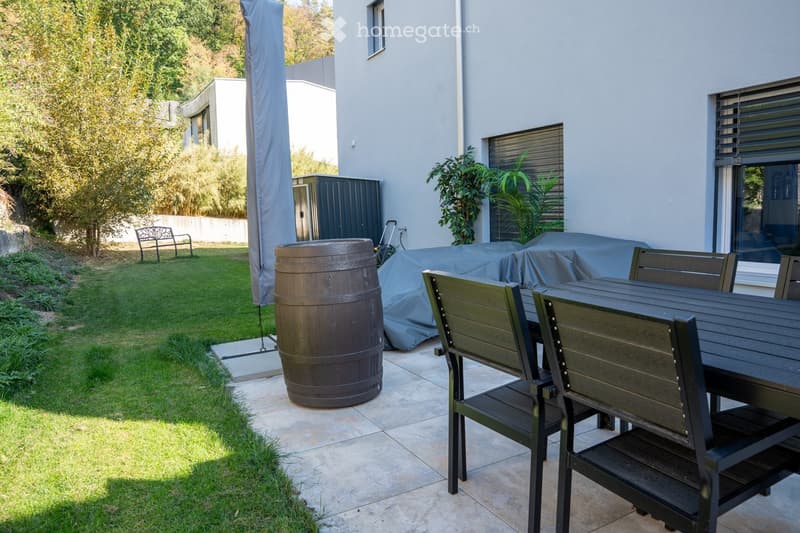 Outdoor patio area with a wooden table and chairs, a large wooden barrel, a covered grill, a bench, and surrounding greenery.