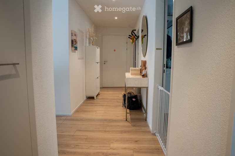 A well-lit hallway with wooden flooring, white walls, and a closed door at the end. Features include a small table with a mirror, a white cabinet, and a framed picture on the wall.