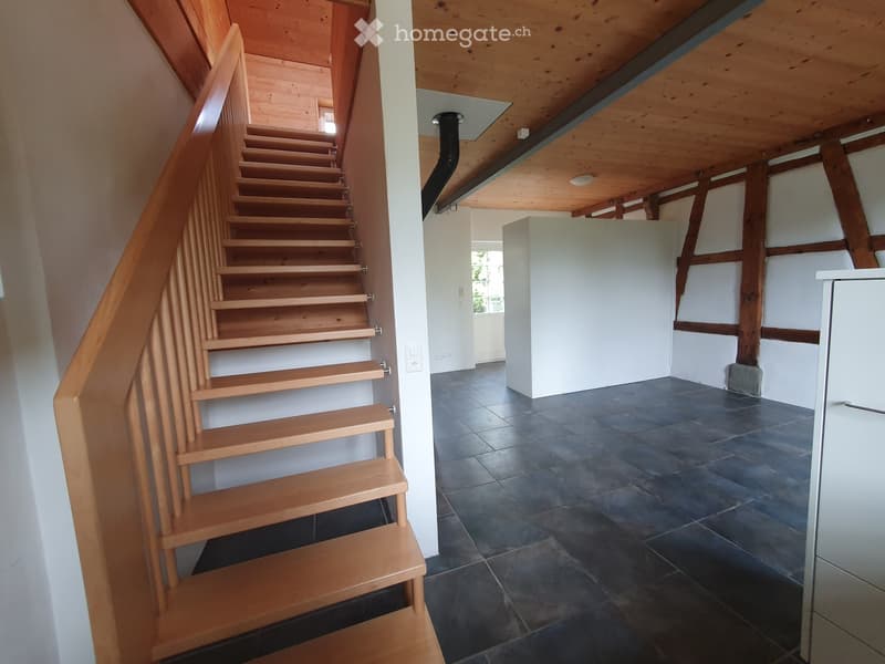 Wooden staircase with a handrail leads to an upper level. The hallway has dark tiled flooring, white walls, and exposed wooden beams on the ceiling.