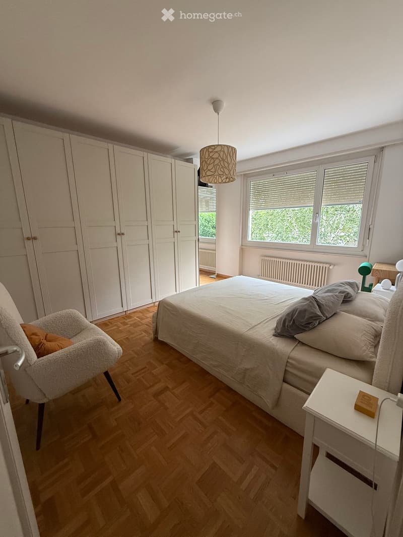 Modern bedroom with wooden flooring, large bed, nightstand, armchair, and built-in wardrobe.