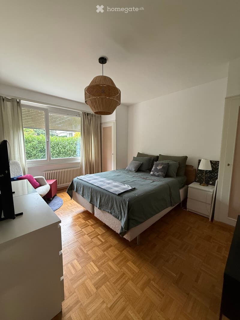Cozy bedroom with wooden flooring, large window with curtains, a double bed with green bedding, a nightstand with a lamp, a small chair, and a table.
