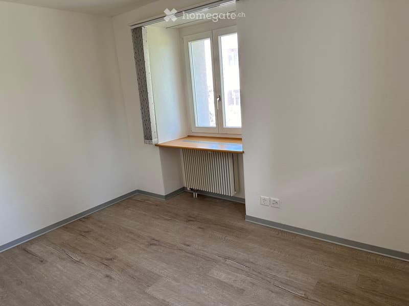Empty room with wooden flooring, white walls, a window with double panes, a radiator, and a small wooden shelf.