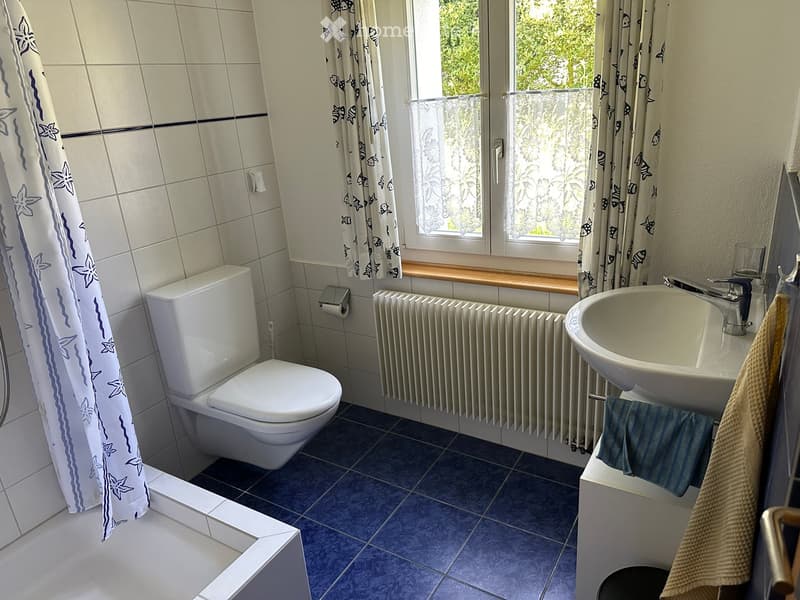 Bathroom with a toilet, sink, radiator, and bath, featuring blue tiles and a window with floral curtains.