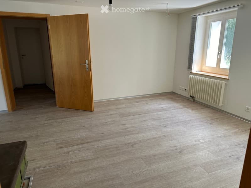 Empty room with wooden flooring, white walls, a window with a radiator, and an open wooden door leading to another space.
