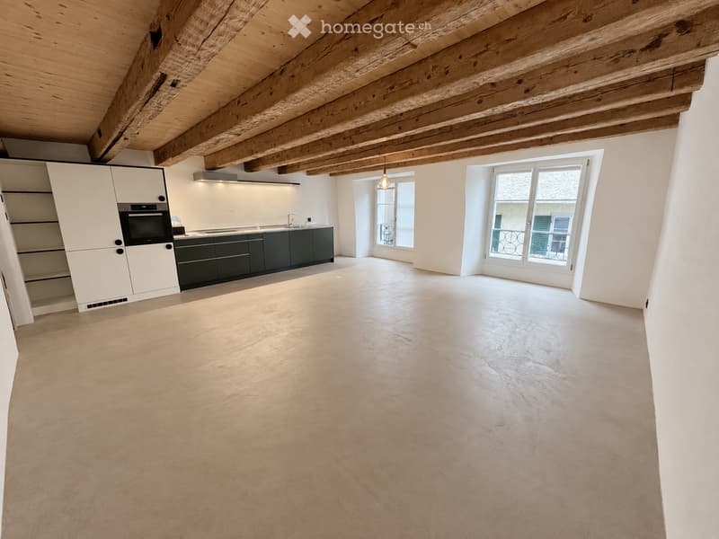 Open, unfurnished room with exposed wooden beams on the ceiling, white walls, large windows, and a modern kitchen area with cabinets and appliances.