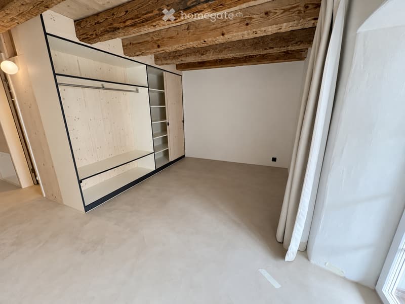 Empty room with exposed wooden beams on the ceiling, white walls, and a built-in closet with glass doors and shelves.