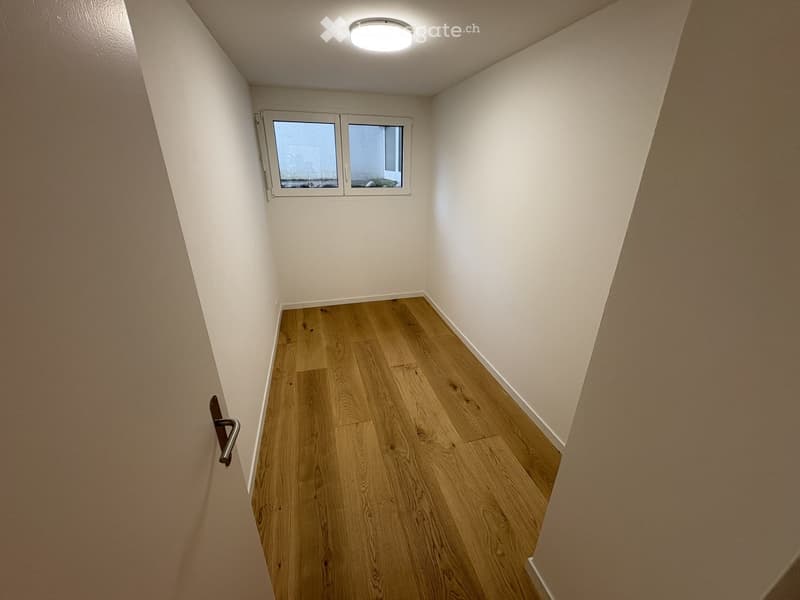 Empty room with wooden flooring, white walls, and a small window at the far end. The room has a simple, clean design with a door on the left side.