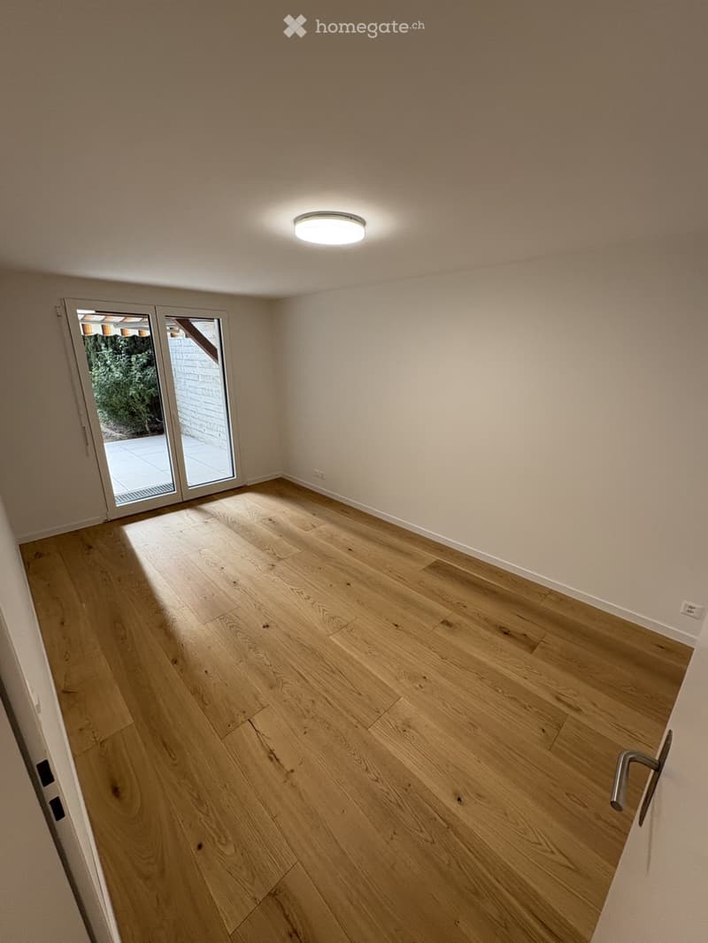 Empty room with light wooden flooring, white walls, and a large sliding glass door leading outside.