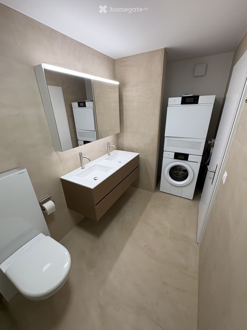 Modern bathroom with a double sink, toilet, mirror, and stacked washer/dryer unit.