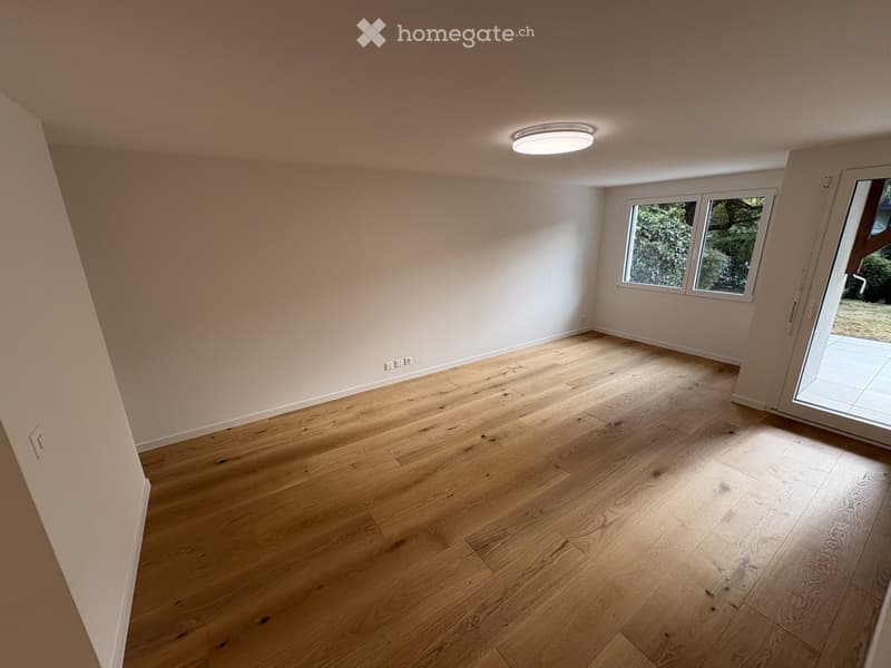 Empty room with light wooden flooring, white walls, and large windows with a sliding glass door leading outside.