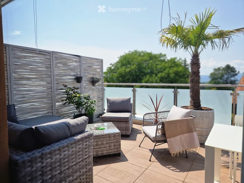 A modern balcony with wicker furniture, including a sofa, chairs, a coffee table, and potted plants, with a view of greenery and a clear blue sky.