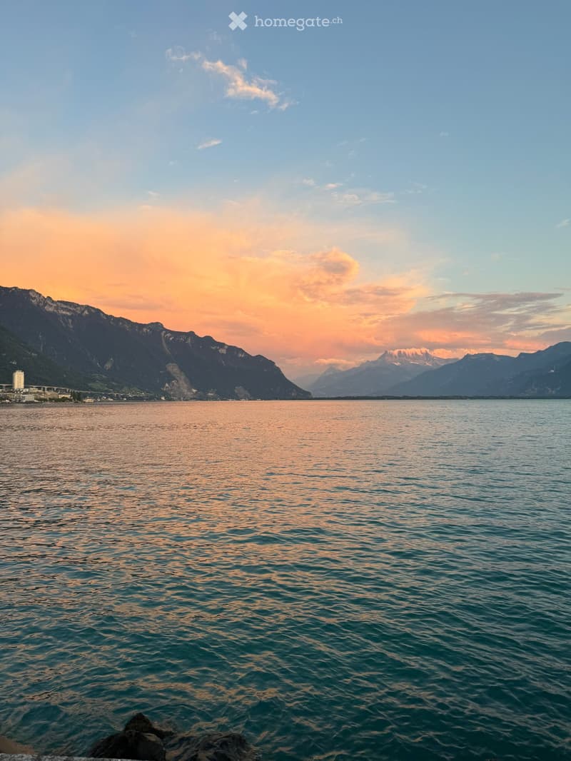 A serene lake view at sunset with calm water reflecting the sky, surrounded by mountains and a distant cityscape.