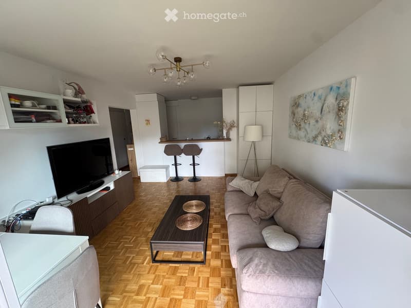 Modern living room with a grey sofa, coffee table, two stools, a TV, a lamp, and a painting on the wall. The room features a wooden herringbone floor and a small kitchen area with bar stools.
