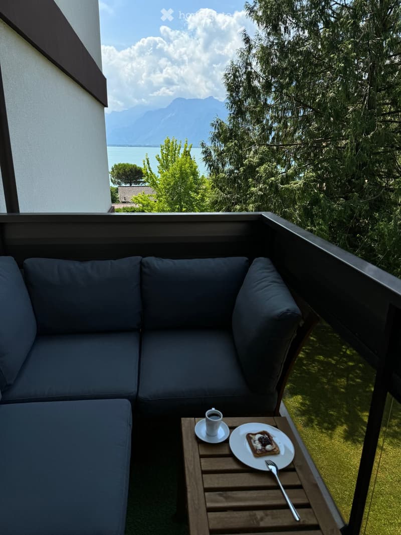 Balcony with a dark blue sofa, a small wooden table holding a cup of coffee, a plate with bread, and a fork. The view shows mountains and a lake in the distance.