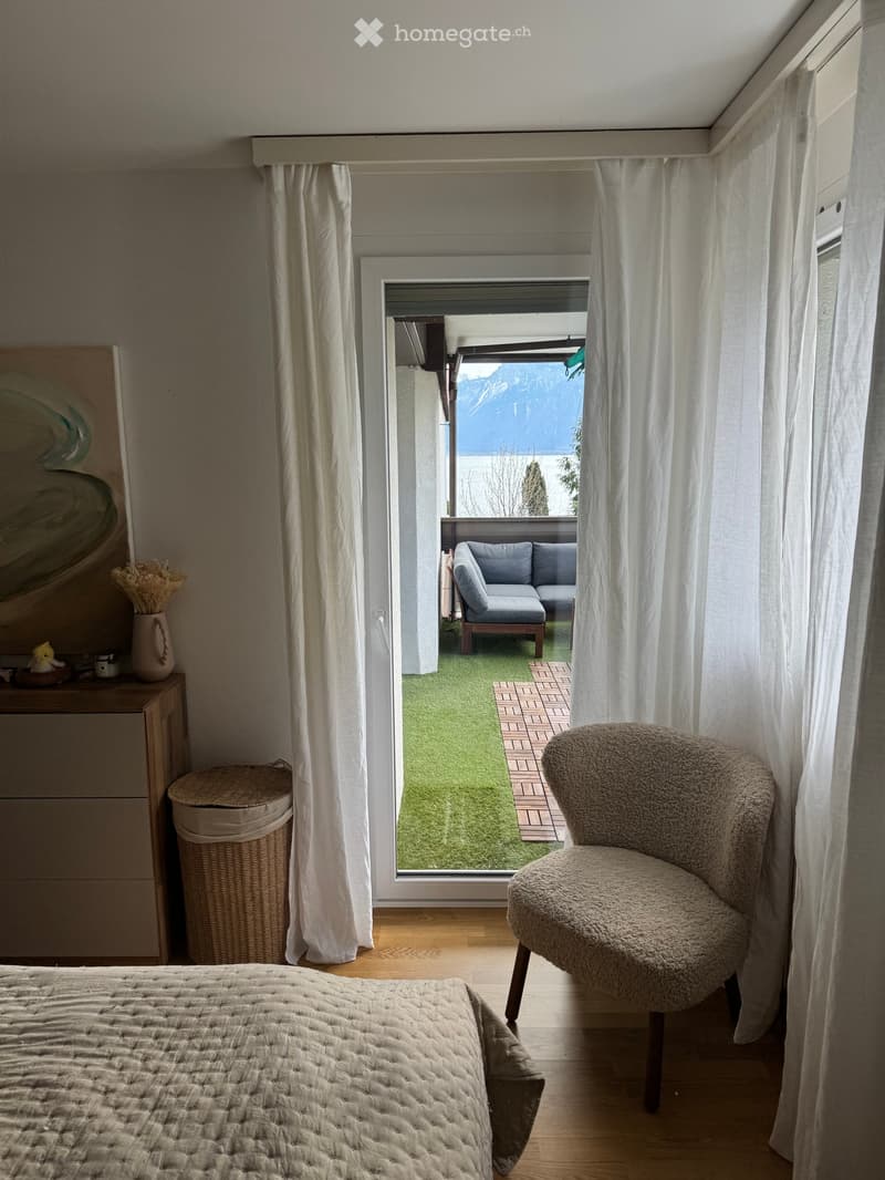 Bright bedroom with wooden flooring, large window with white curtains, bed, armchair, small table with vase and decorative items, and a view of a balcony with artificial grass and a blue sofa.