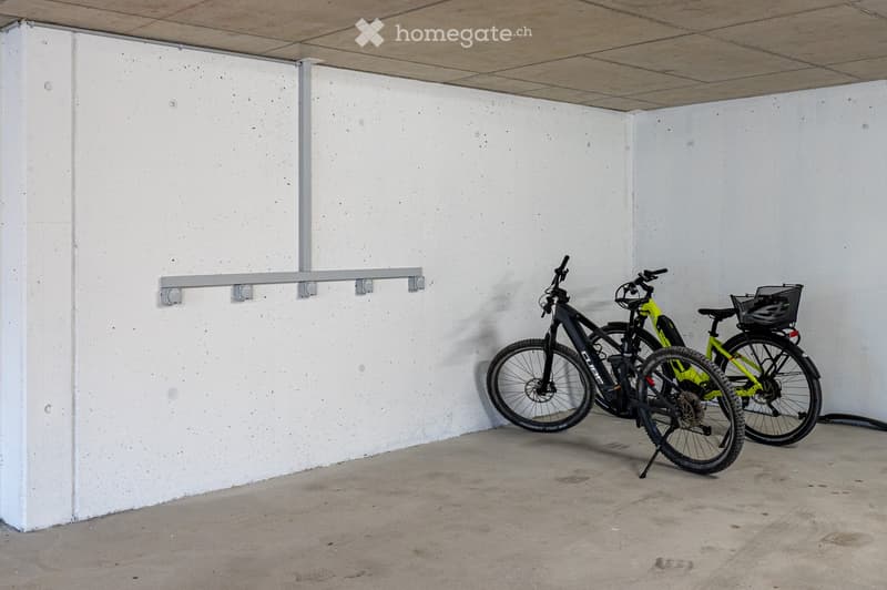 Two bicycles, one black and one green, are parked in a concrete-walled area with a metal bike rack attached to the wall.