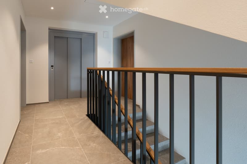Modern hallway with tiled floor, white walls, and a staircase with wooden steps and metal railing. Two doors are visible, one grey and one wooden.