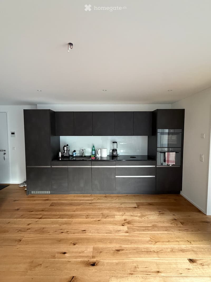 Modern kitchen with dark grey cabinets, wooden flooring, and stainless steel appliances including a sink, coffee maker, and blender. The space is well-lit with recessed ceiling lights.