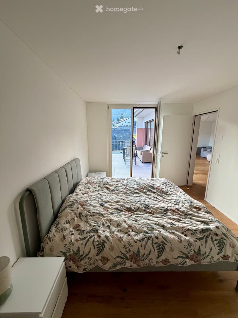 Bedroom with a large bed, floral duvet, nightstand, and an open sliding door leading to a balcony.