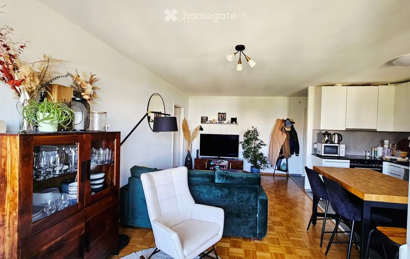 A modern living room with a teal sofa, white armchair, wooden cabinet, TV, and a small kitchen area with a table and stools.