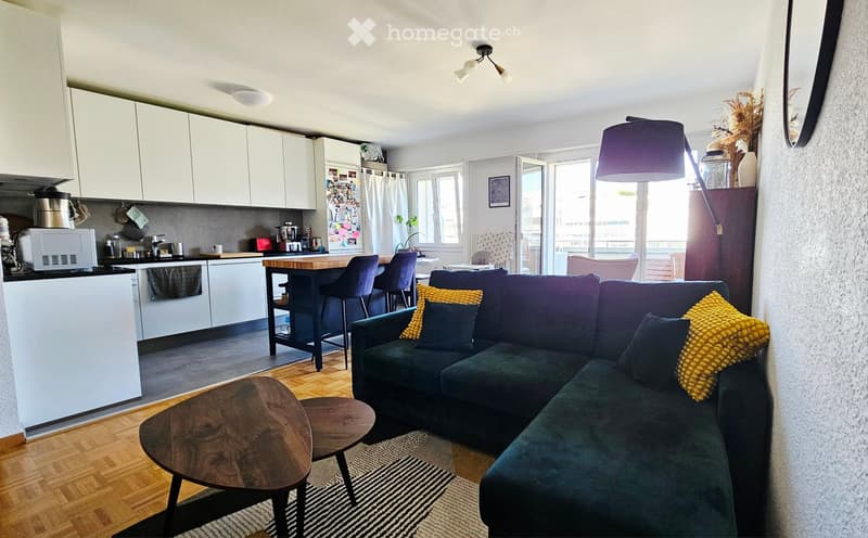 Open-plan living and dining area with a green sectional sofa, wooden coffee table, bar stools, and a modern kitchen with white cabinets and stainless steel appliances.