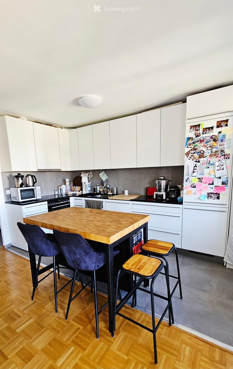 Modern kitchen with white cabinets, a wooden island, and various appliances including a refrigerator, microwave, and stove. There are two blue chairs and two wooden stools around the island.