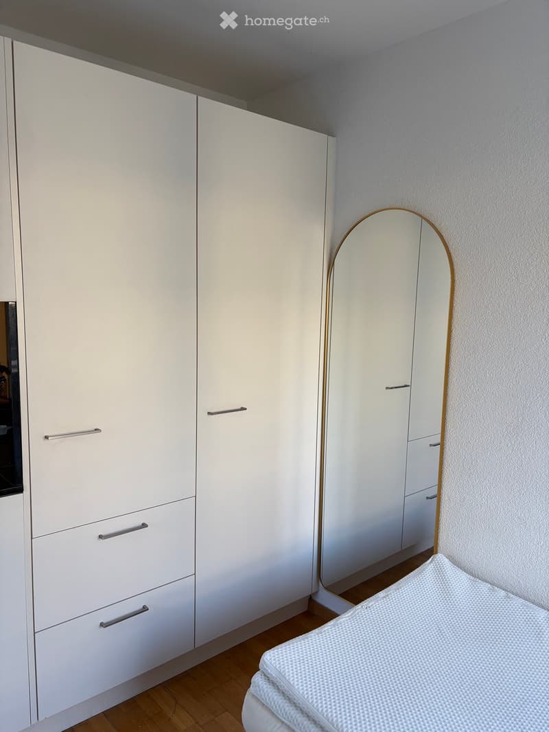 Modern bedroom with white built-in wardrobe, a round mirror with a gold frame, and a neatly made bed with white bedding.