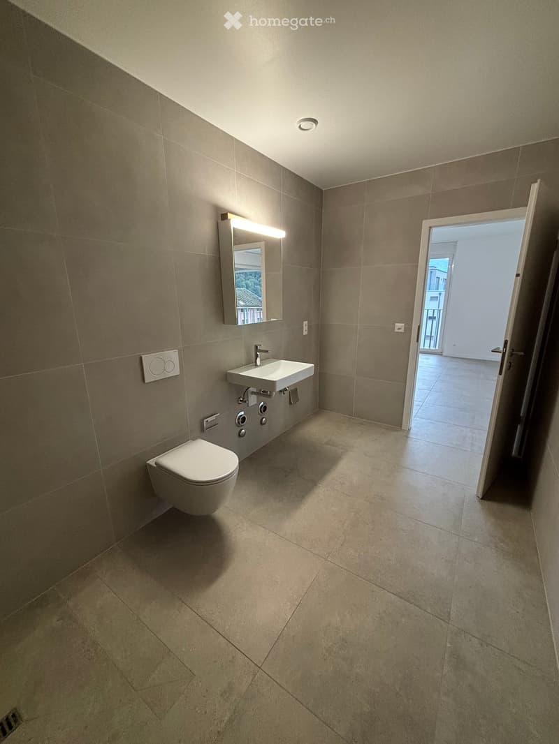 Modern bathroom with beige tiles, a toilet, a sink with a mirror, and an open doorway leading to another room.