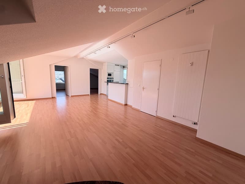 Empty room with wooden flooring, white walls, and a small kitchen area visible in the background. No furniture or personal items are present.